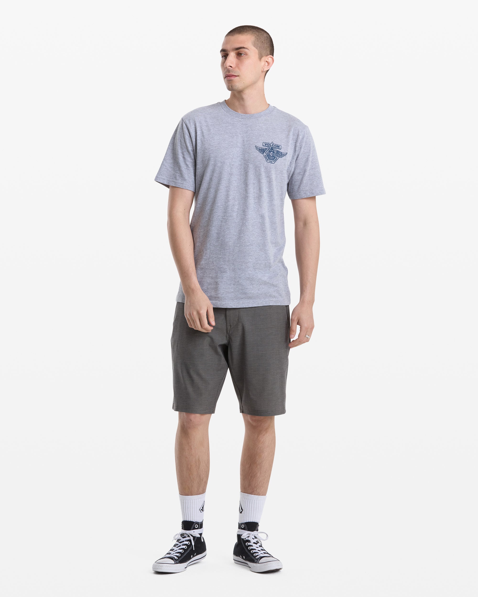 A man faces slightly to the side in a Volcom Men's Free Wheel Short Sleeve Tee (light gray), dark gray shorts, white crew socks, and black sneakers against a plain white background.