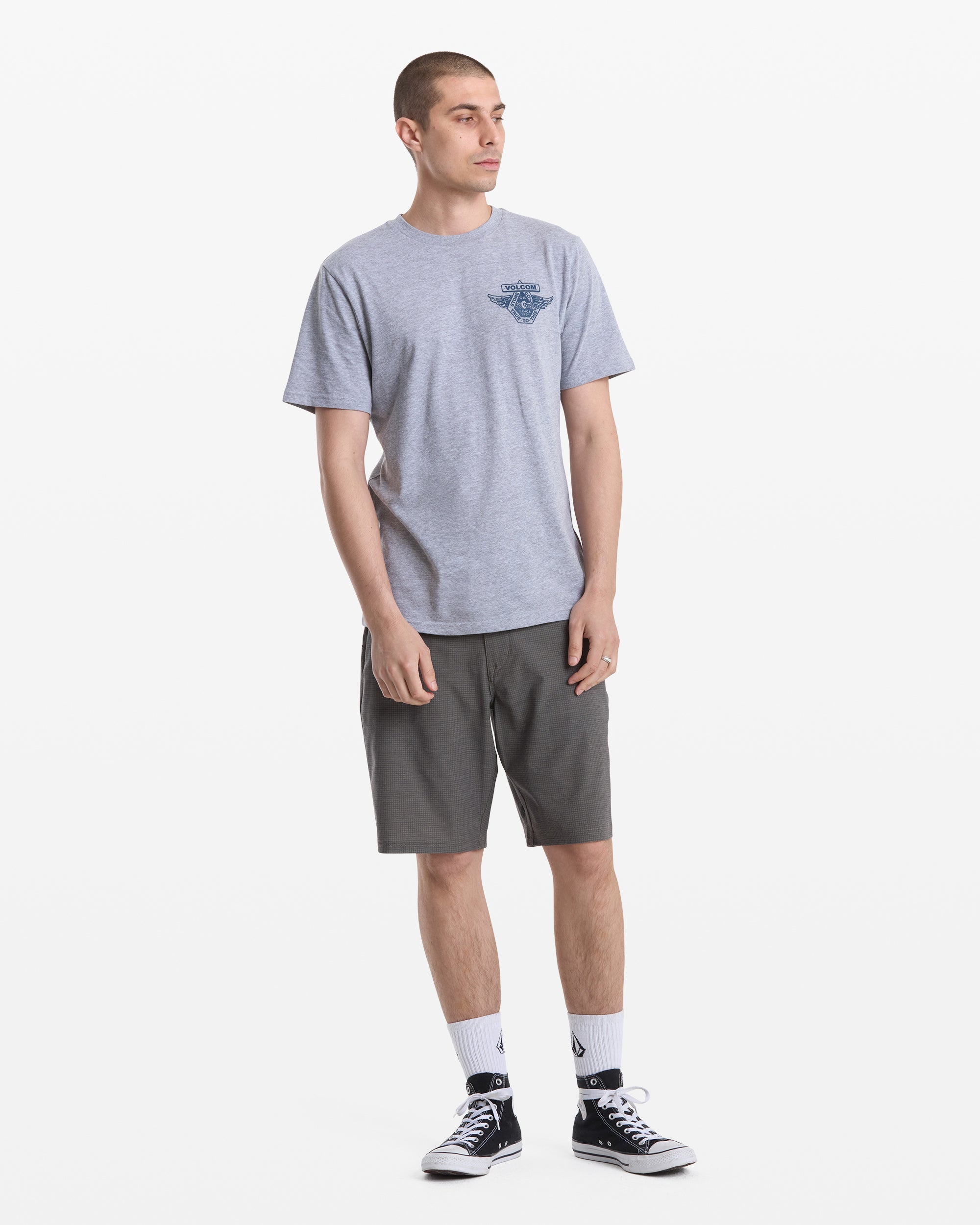 A young man with short hair stands against a white background wearing the Volcom Men's Free Wheel Short Sleeve Tee, paired with dark gray shorts, white crew socks, and black sneakers. He looks slightly right with his hands by his sides.