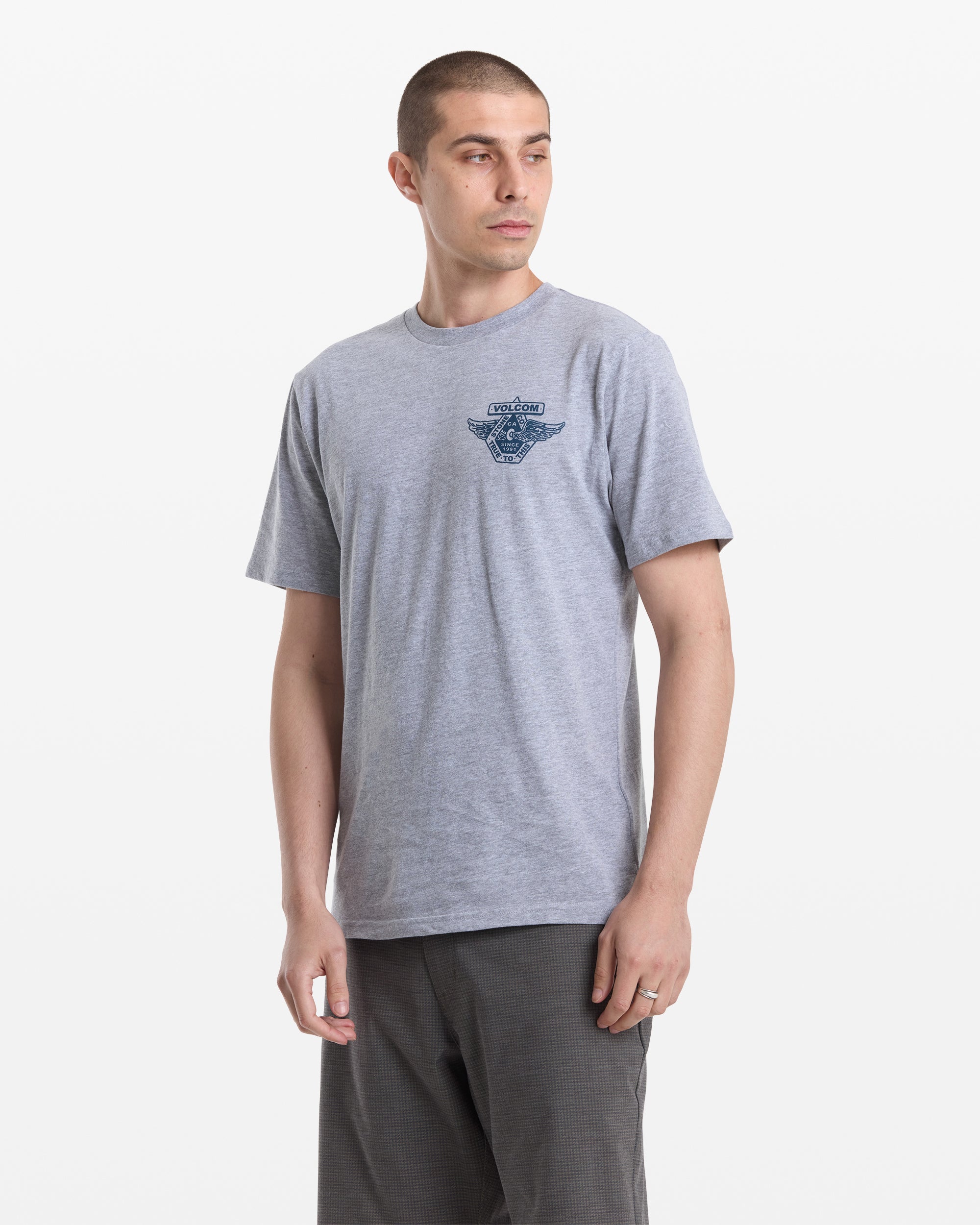 A man with short hair wears the Volcom Men's Free Wheel Short Sleeve Tee in light gray, paired with dark gray shorts, standing against a plain white background and looking slightly to his left.
