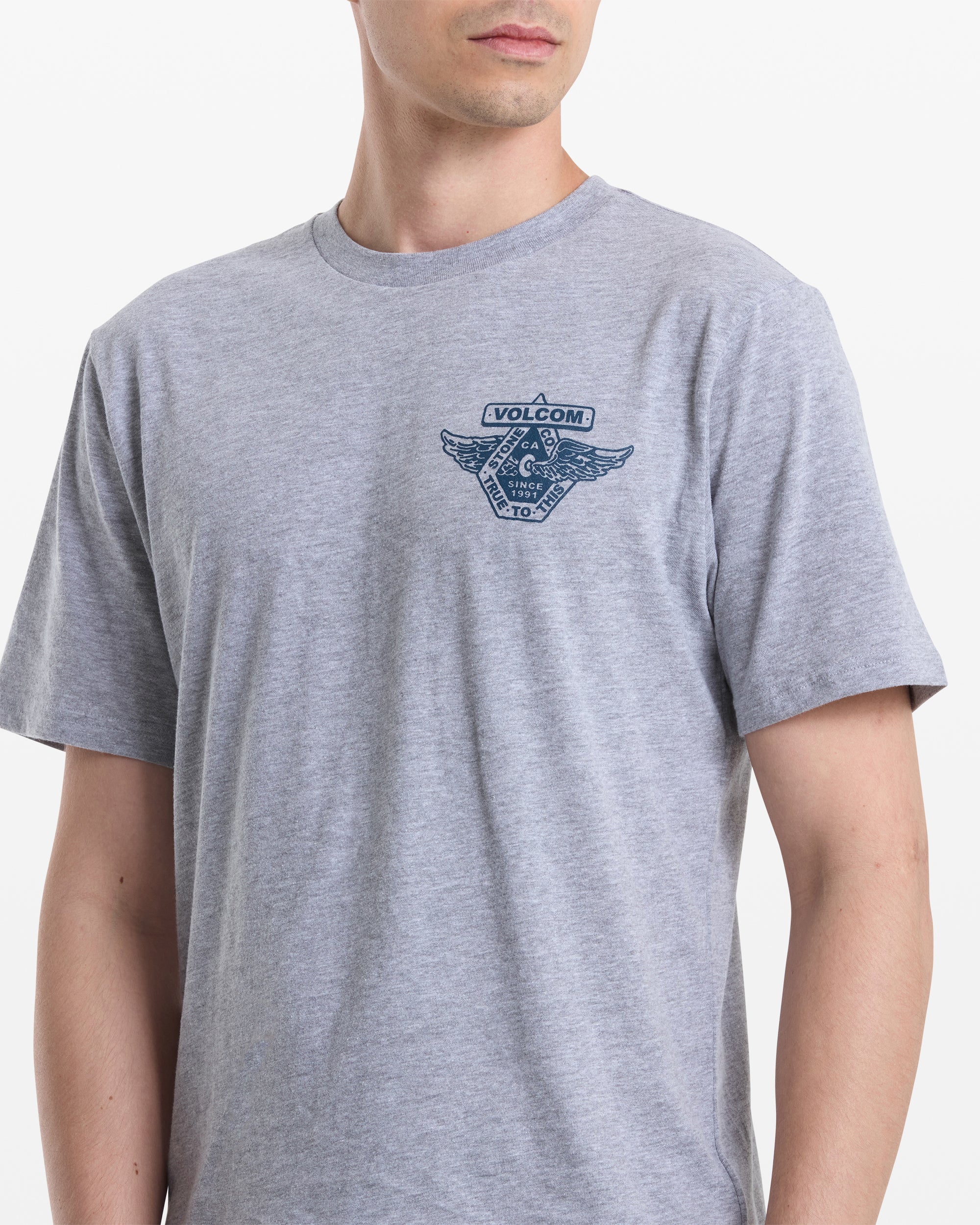 A man wears the Volcom Men's Free Wheel Short Sleeve Tee in light grey, featuring a small blue logo with wings and geometric shapes on the left chest. He faces forward with his head slightly turned to the side.