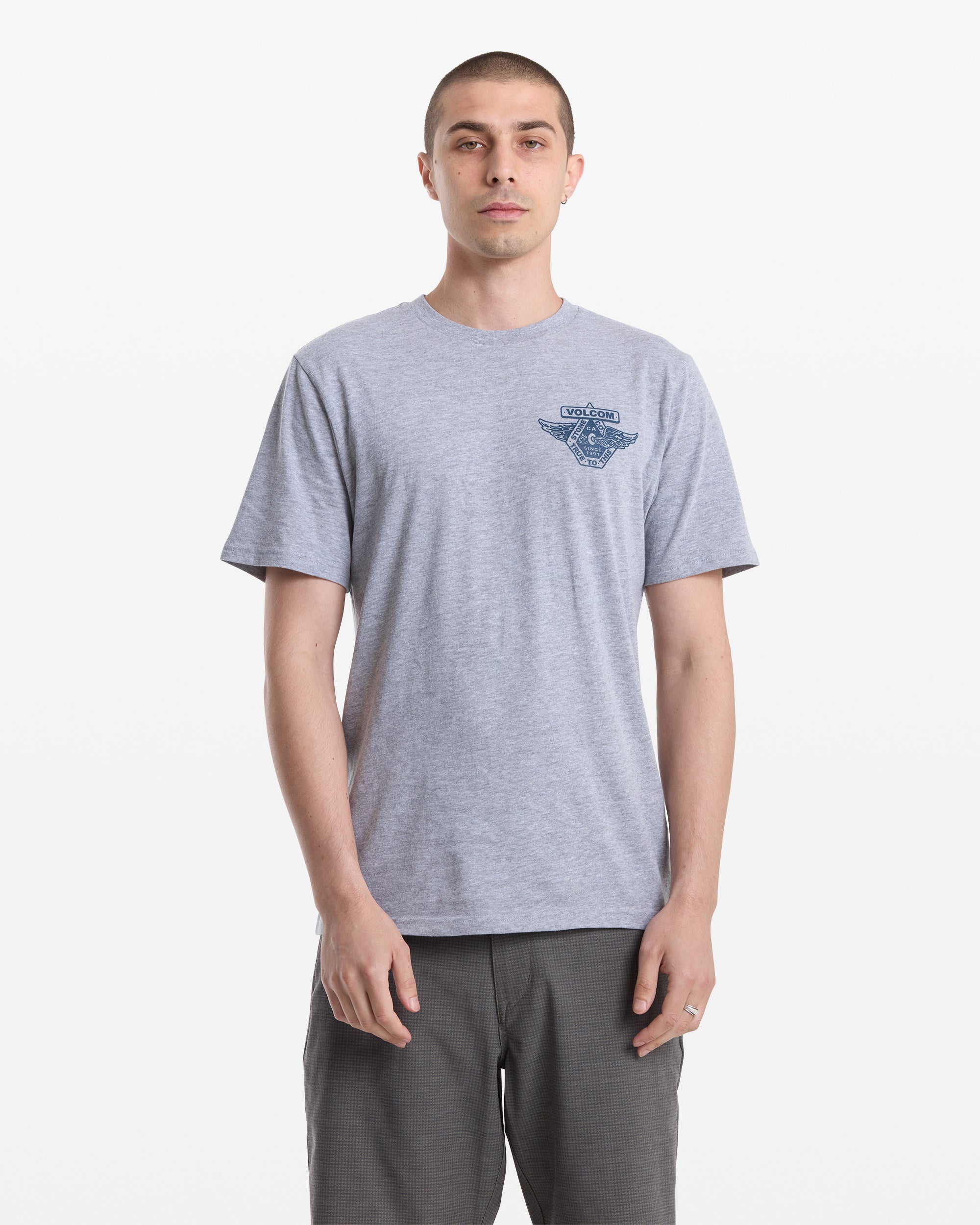 A man with short hair wears the Volcom Men's Free Wheel Short Sleeve Tee in light gray, featuring a small dark blue graphic and text on the left chest, paired with dark pants, standing against a plain white background.