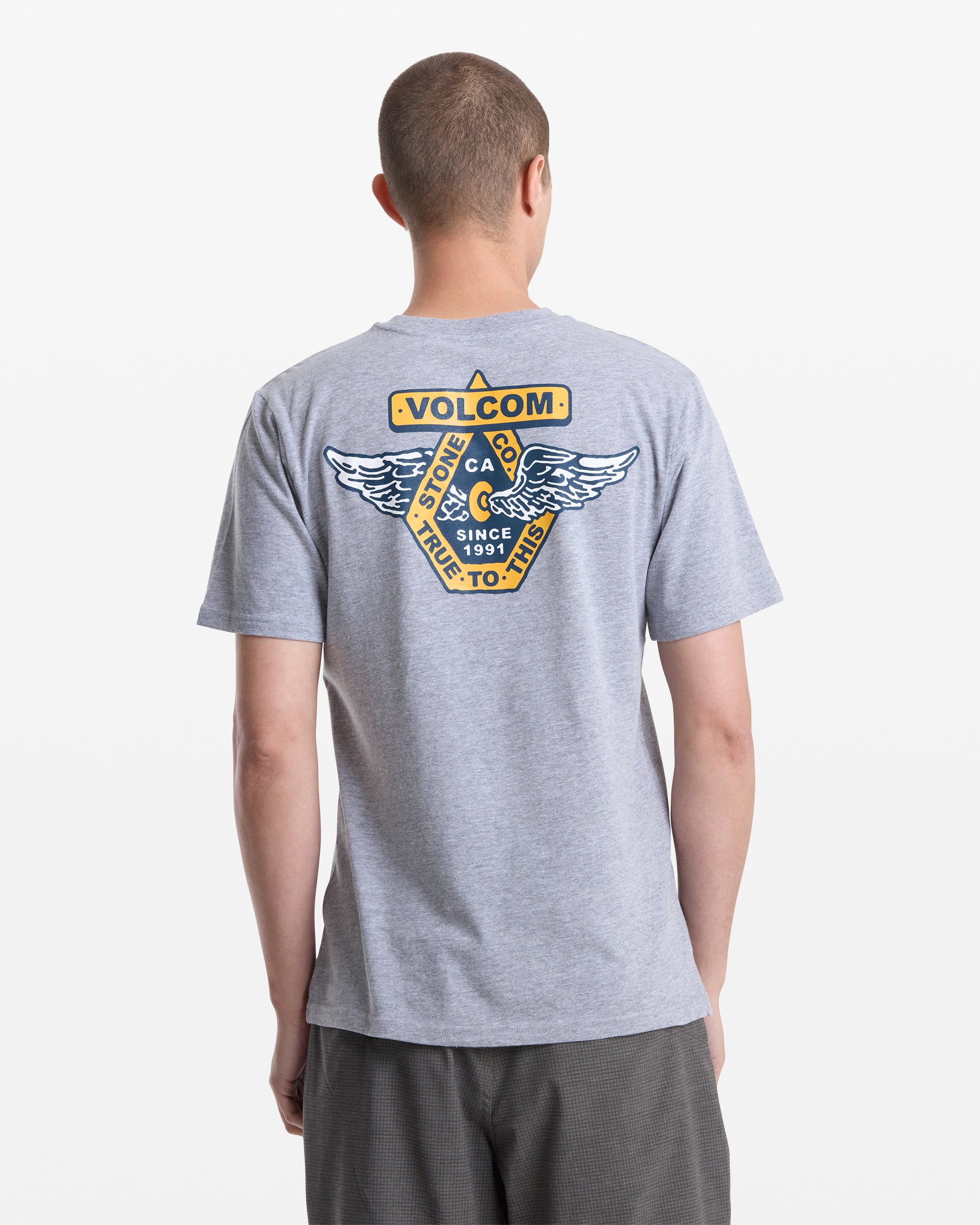 A person is seen from the back wearing the Volcom Men's Free Wheel Short Sleeve Tee in gray, featuring a large yellow and blue winged emblem with "Volcom," "True To This," and "Since 1991" printed on it.