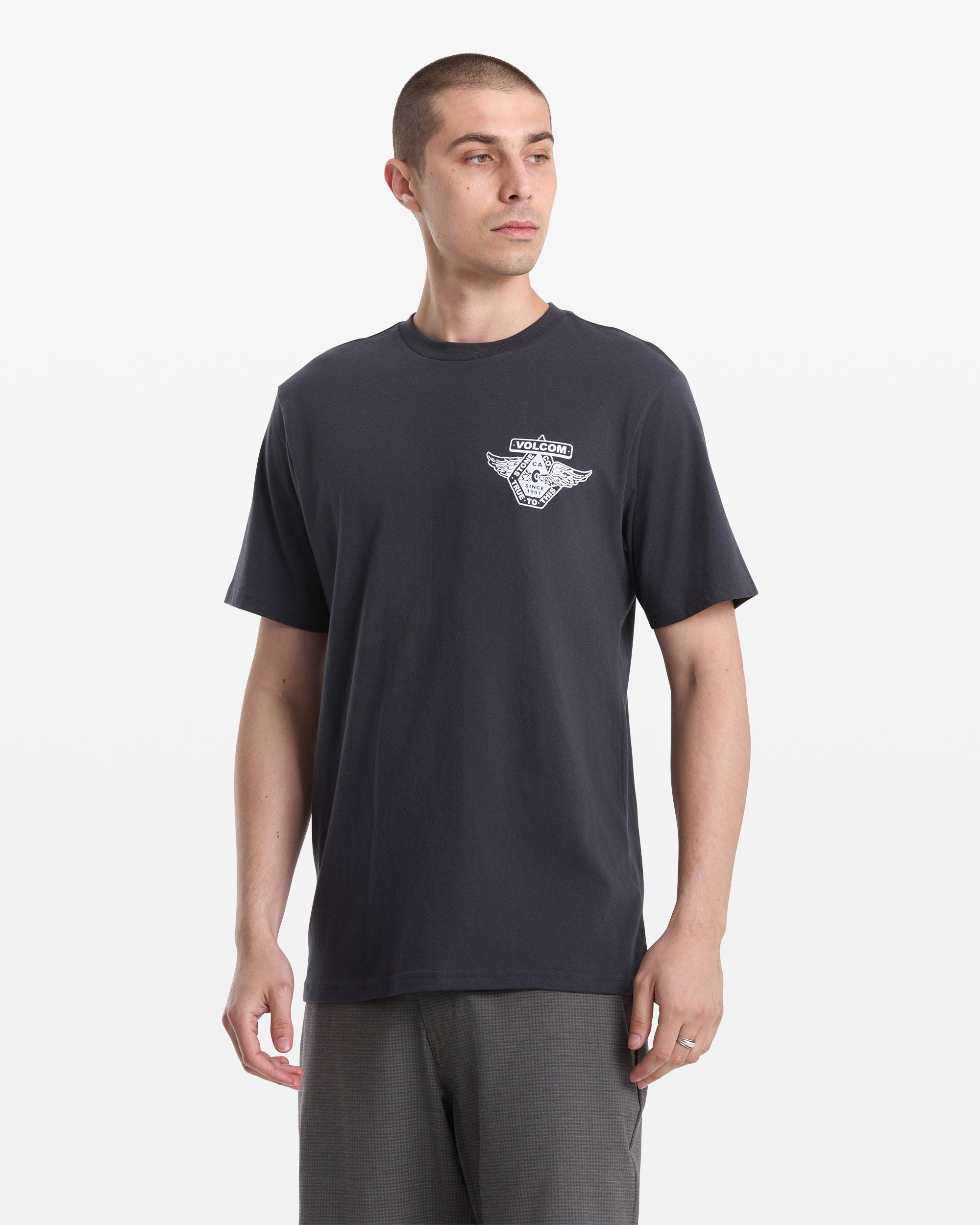 A man with short hair models the Volcom Men's Free Wheel Short Sleeve Tee, featuring a small left chest graphic, paired with gray pants and standing against a plain white background.
