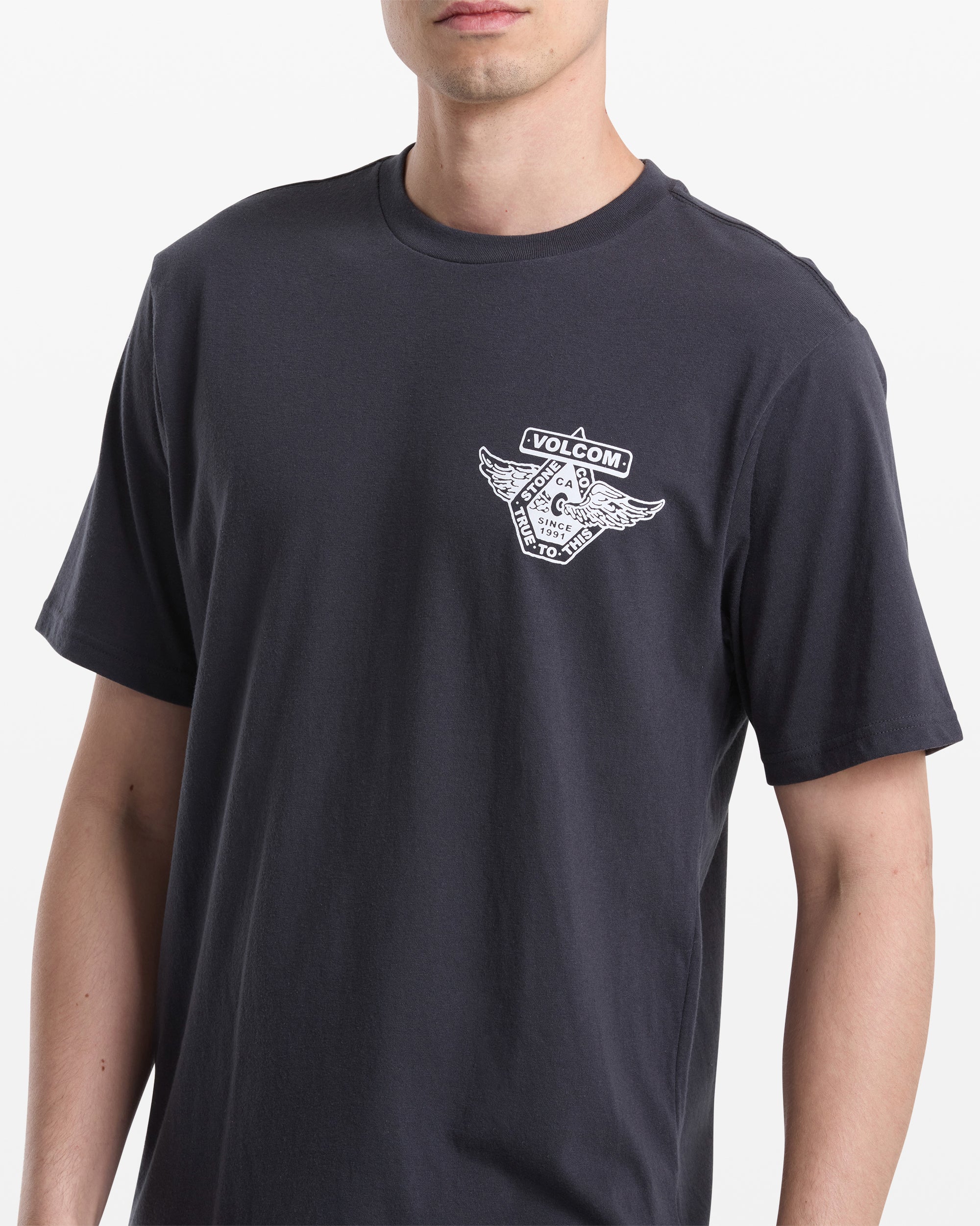 A person wears the Volcom Men's Free Wheel Short Sleeve Tee, a plain black T-shirt featuring a small white triangle and wings graphic with Volcom text on the left chest. The background is plain white.