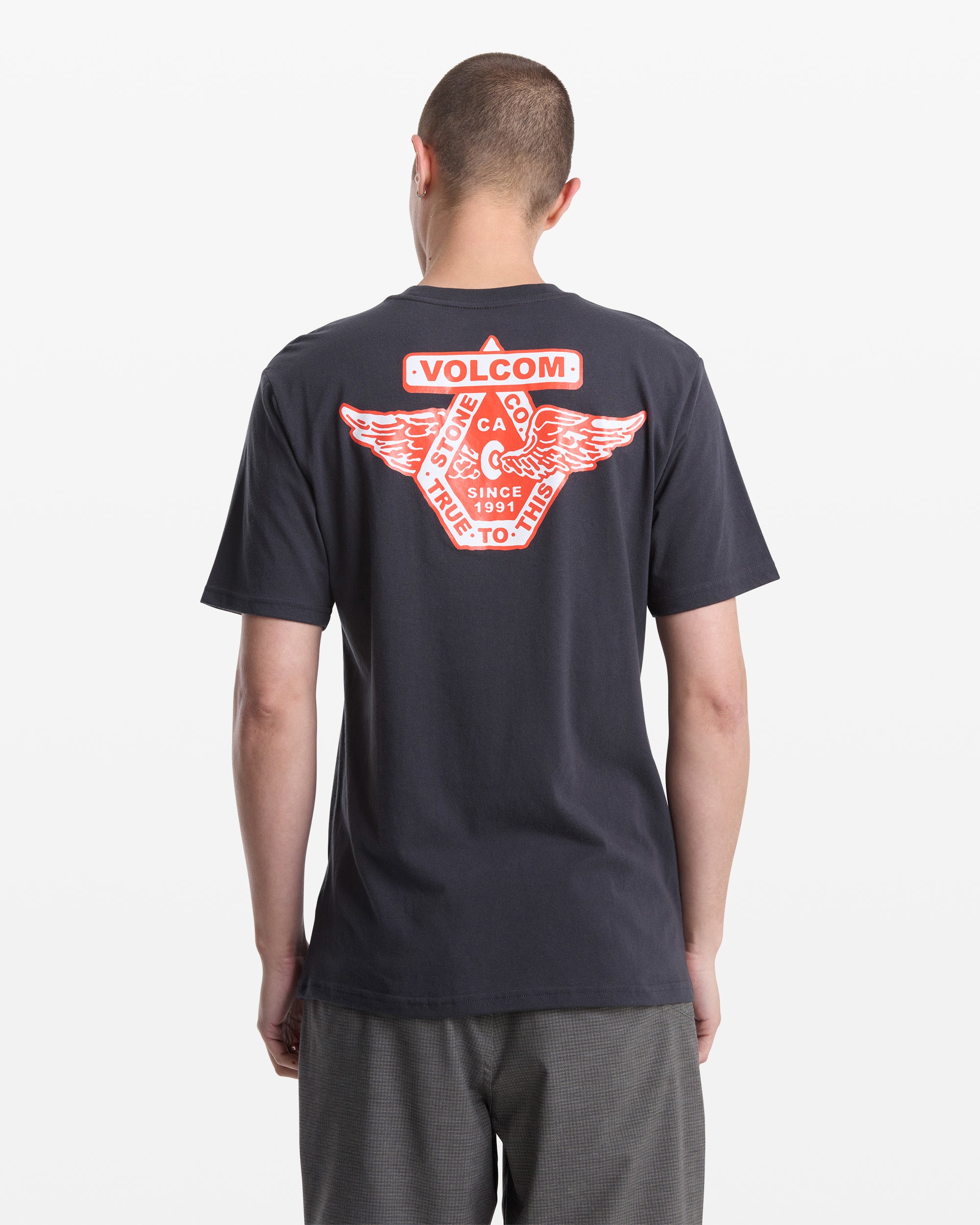 A man wears the Volcom Men's Free Wheel Short Sleeve Tee, featuring a large red and white winged graphic and text on the back, set against a plain white background.