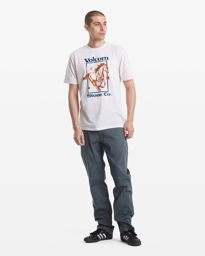 A young man with short hair wears a white Volcom Men's Giddy Up Short Sleeve Tee, gray pants, and black Adidas sneakers. He stands facing slightly to the left against a plain white background.