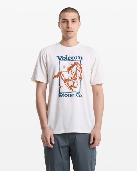 A man with short hair wears the Volcom Men's Giddy Up Short Sleeve Tee, which features a horse and stars graphic. He faces forward with a neutral expression, wearing gray pants against a plain white background.