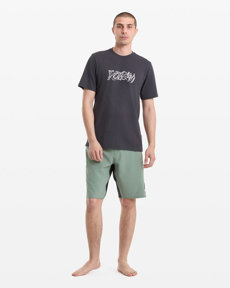 A young man stands barefoot against a white background, wearing the Volcom Men's Destroyed Short Sleeve Tee in black with white lettering and light green shorts. He faces forward with a neutral expression.