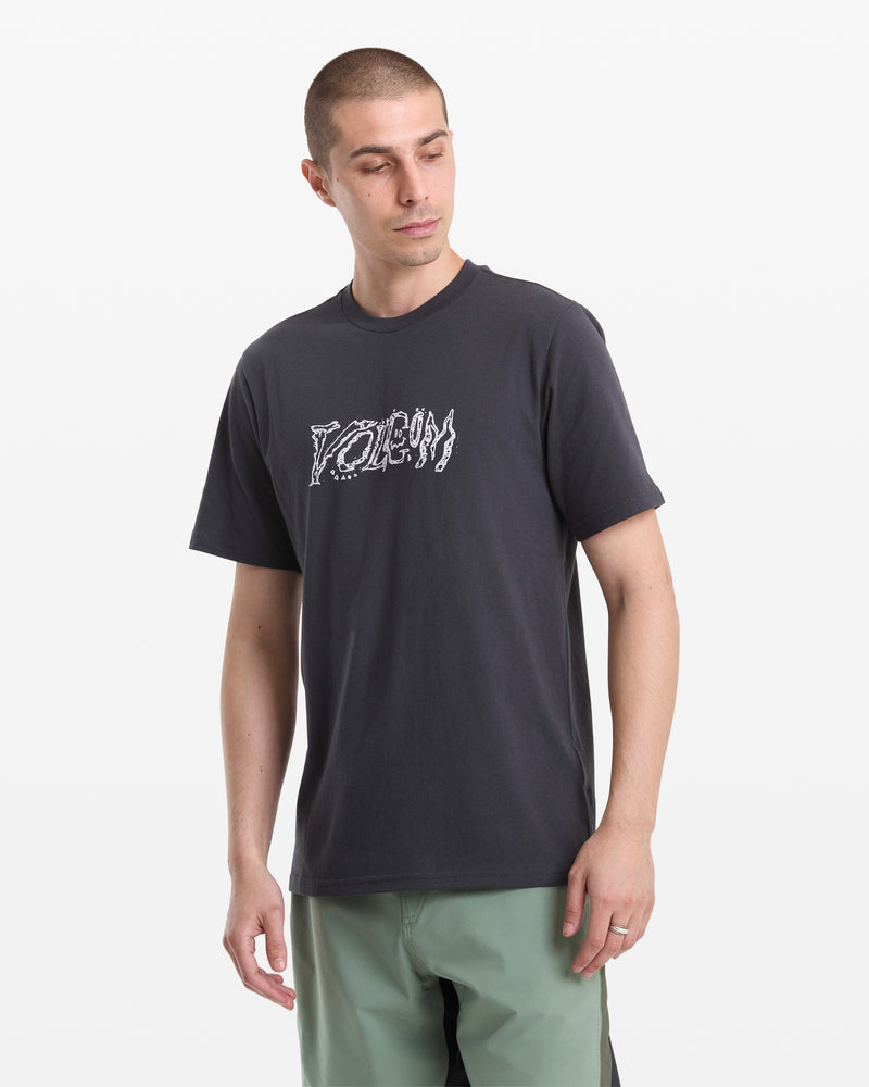 A man with short hair wears the Volcom Men's Destroyed Short Sleeve Tee, a dark t-shirt with a stylized white POEM graphic on the chest, and light green shorts, standing against a plain white background.