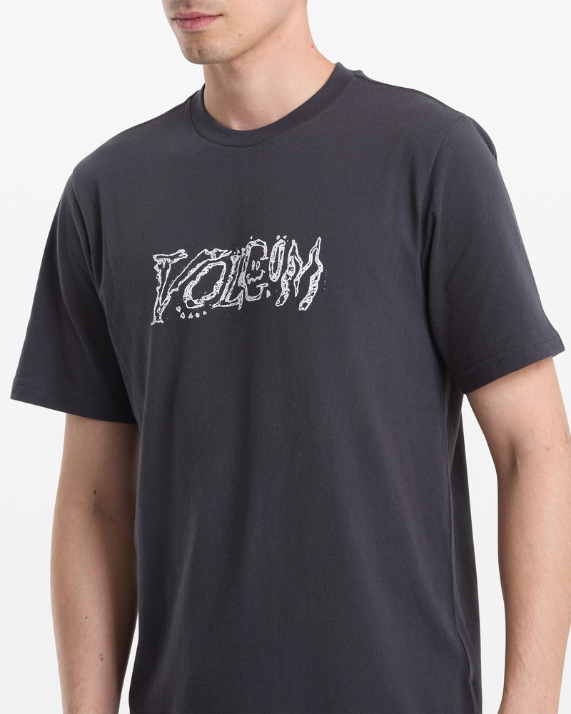 A person wearing the Volcom Men's Destroyed Short Sleeve Tee, featuring a bold, jagged white "VOLCOM" logo on a dark shirt. Their head is partially out of frame against a plain white background.