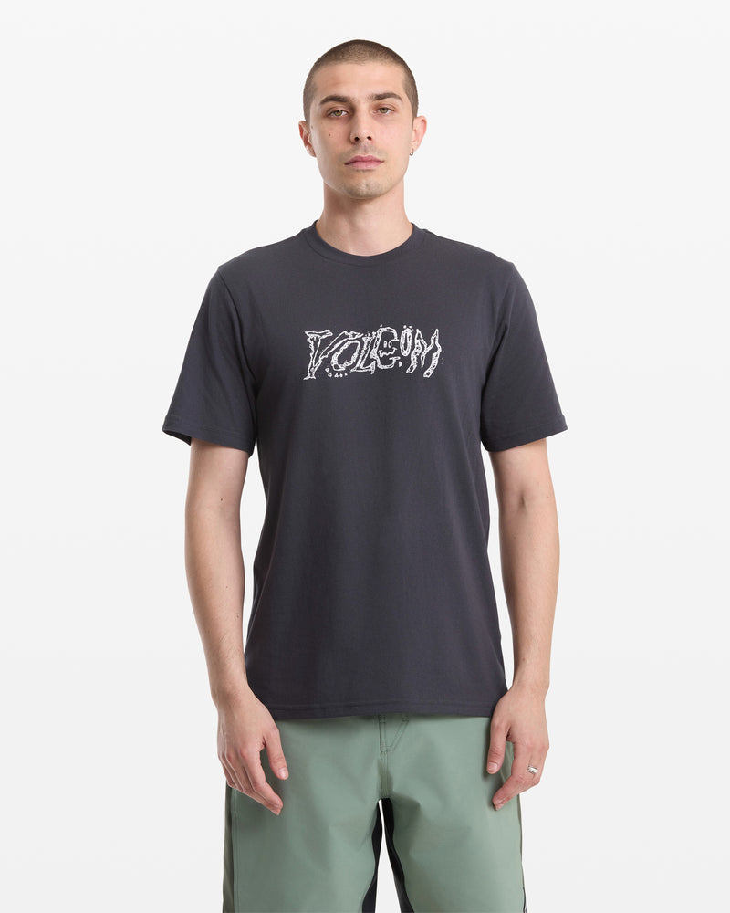A man with short hair wears the Volcom Men's Destroyed Short Sleeve Tee, featuring POISON in a melting white graphic font, paired with light green shorts. He stands facing forward against a plain white background.