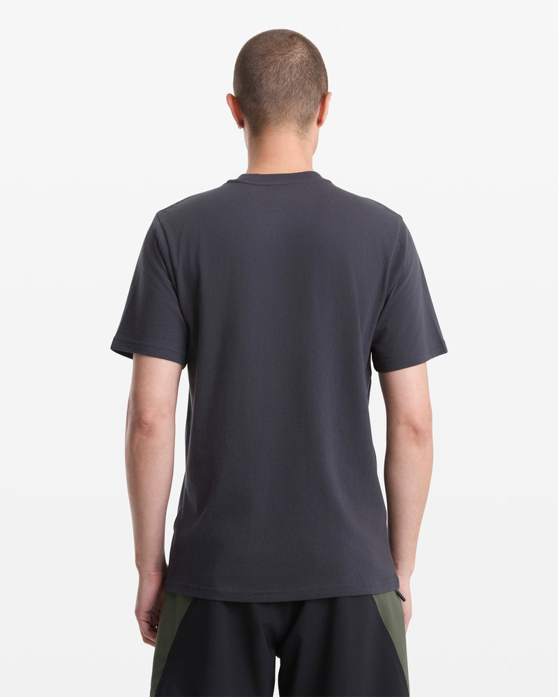 A person with short hair is facing away from the camera, wearing a Volcom Men's Destroyed Short Sleeve Tee in plain dark gray and dark shorts with olive green panels, set against a white background.