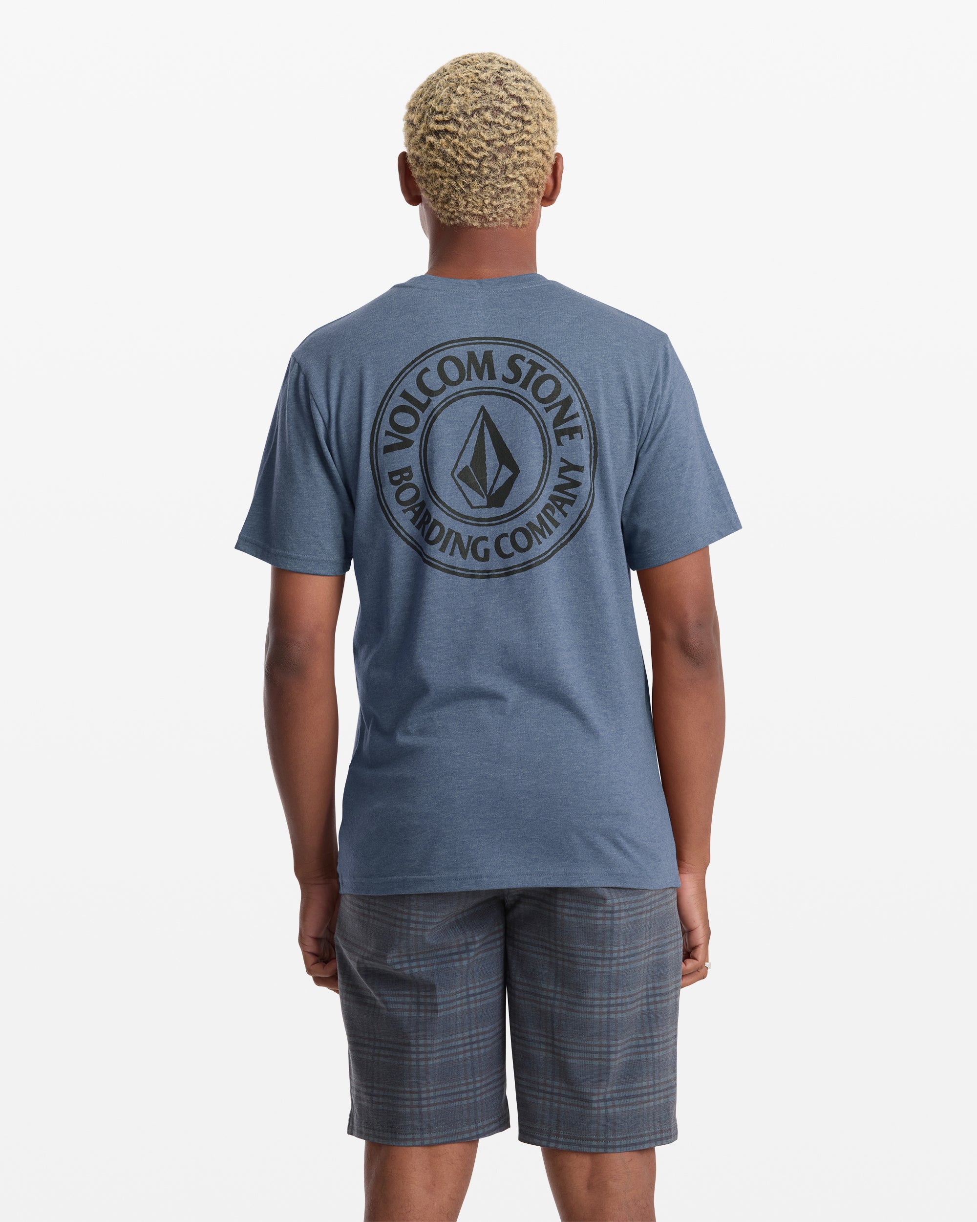 A person with short, curly blond hair is seen from behind wearing a blue Volcom Men's Drain You Short Sleeve Tee with a large circular Volcom logo and text on the back, paired with plaid shorts.