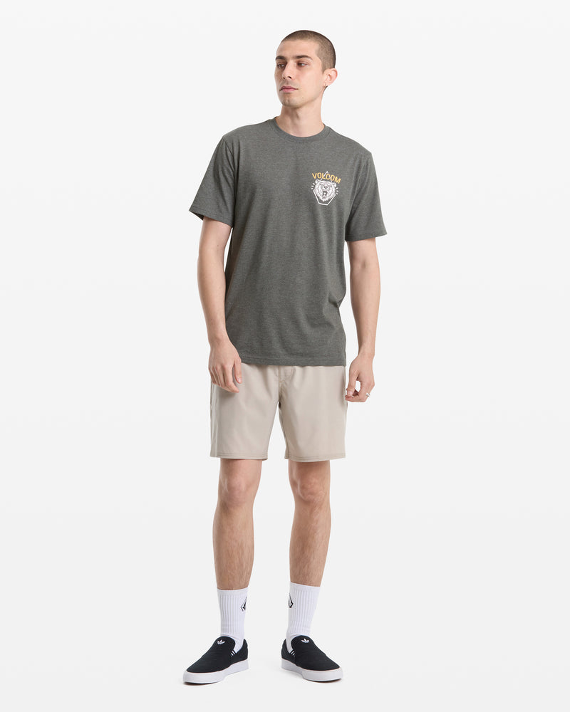 A young man stands against a white background wearing the Volcom Men's Golden Statement Short Sleeve Tee in gray, paired with beige shorts, white crew socks, and black slip-on shoes. He gazes slightly to the side with his arms relaxed at his sides.