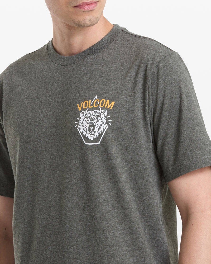 A person wearing the Volcom Men's Golden Statement Short Sleeve Tee in dark gray, featuring a white lion’s head graphic with orange VOLCOM text above it on the left chest.