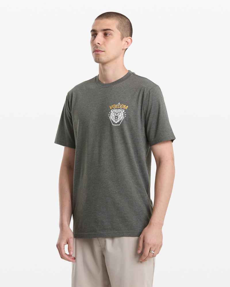 A man with short hair wears the Volcom Men's Golden Statement Short Sleeve Tee, a dark gray shirt featuring a small graphic and logo on the left chest, while standing against a plain white background and looking slightly to the side.