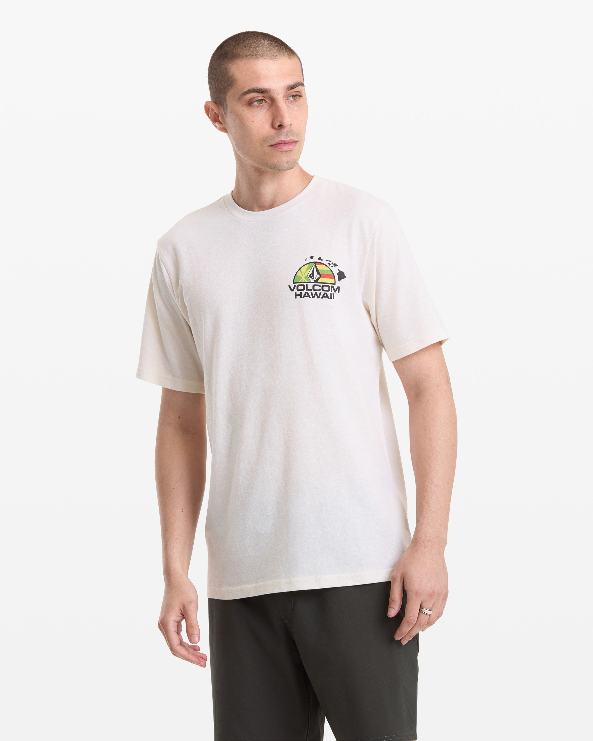 A man stands against a white background, looking slightly left, wearing the Volcom Men's Allegiance Short Sleeve Tee—a light-colored T-shirt with a small, colorful Hawaii graphic on the chest—paired with dark shorts and a simple ring.