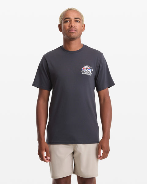 A man with short blond hair wears the Volcom Men's Allegiance Short Sleeve Tee, a dark shirt with a small graphic and HAWAII text on the left chest, along with light-colored shorts, standing against a plain white background.