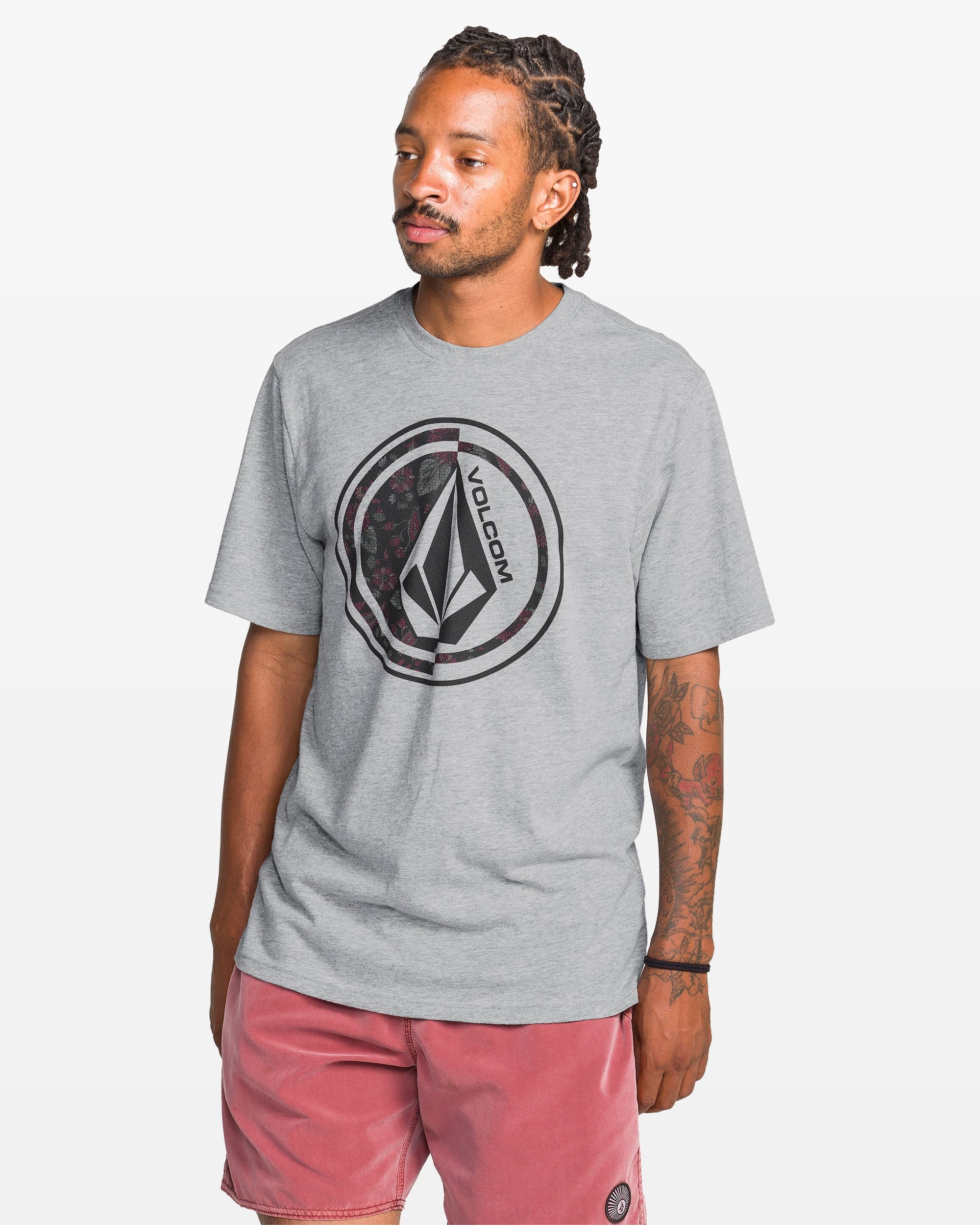 A man with braided hair and tattoos on his left arm wears the Volcom Men's Coop Short Sleeve Tee in light gray, featuring a large logo. Made from a cotton-poly blend, it's paired with pink shorts against a plain white background as he looks slightly left.