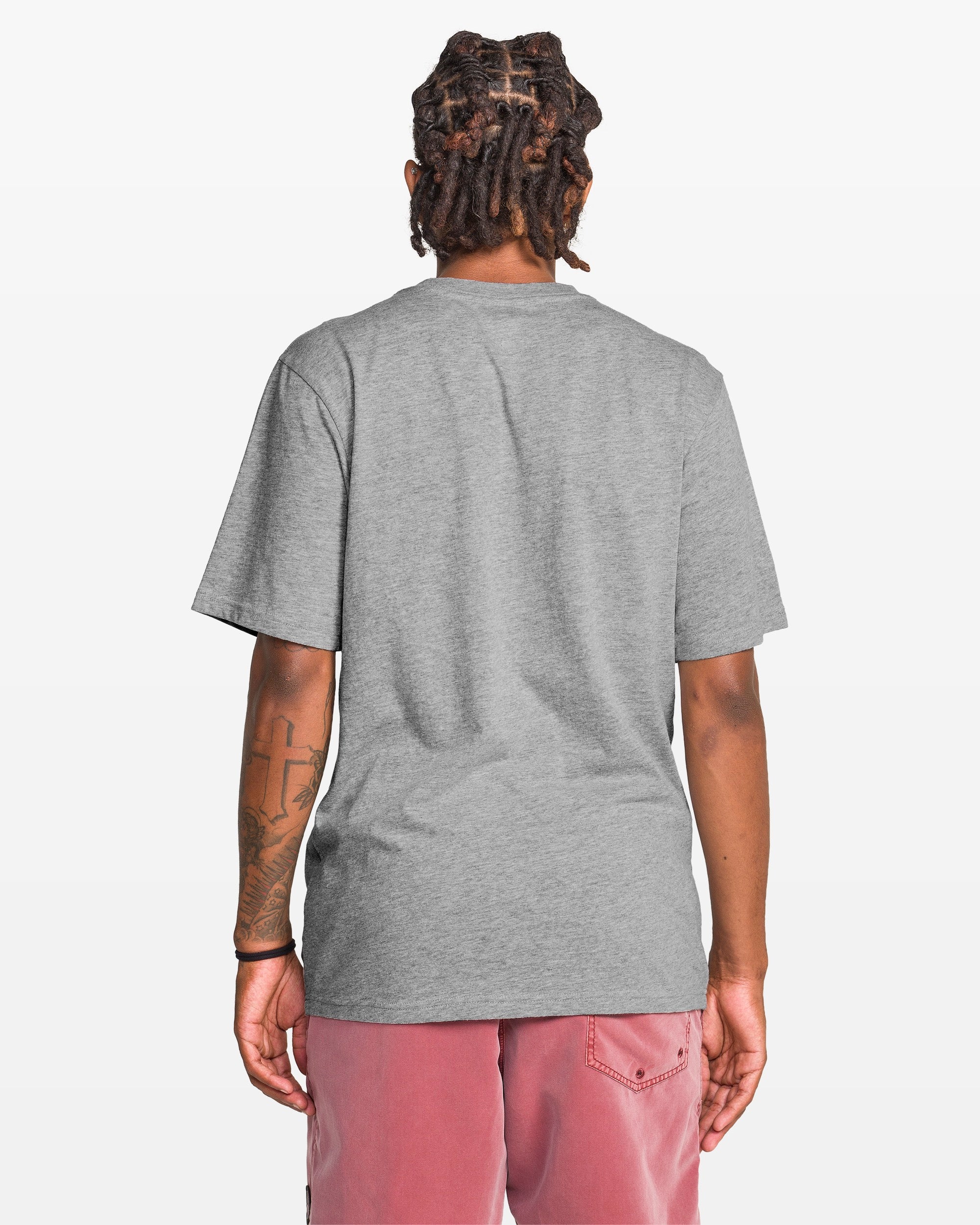 A person with short dreadlocks is shown from behind, wearing the Volcom Men's Coop Short Sleeve Tee in plain gray and faded pink shorts. Their left arm has a tattoo, and the background is white.
