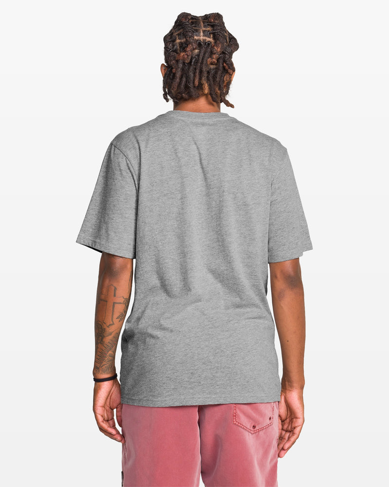 A person with short dreadlocks is shown from behind, wearing the Volcom Men's Coop Short Sleeve Tee in plain gray and faded pink shorts. Their left arm has a tattoo, and the background is white.