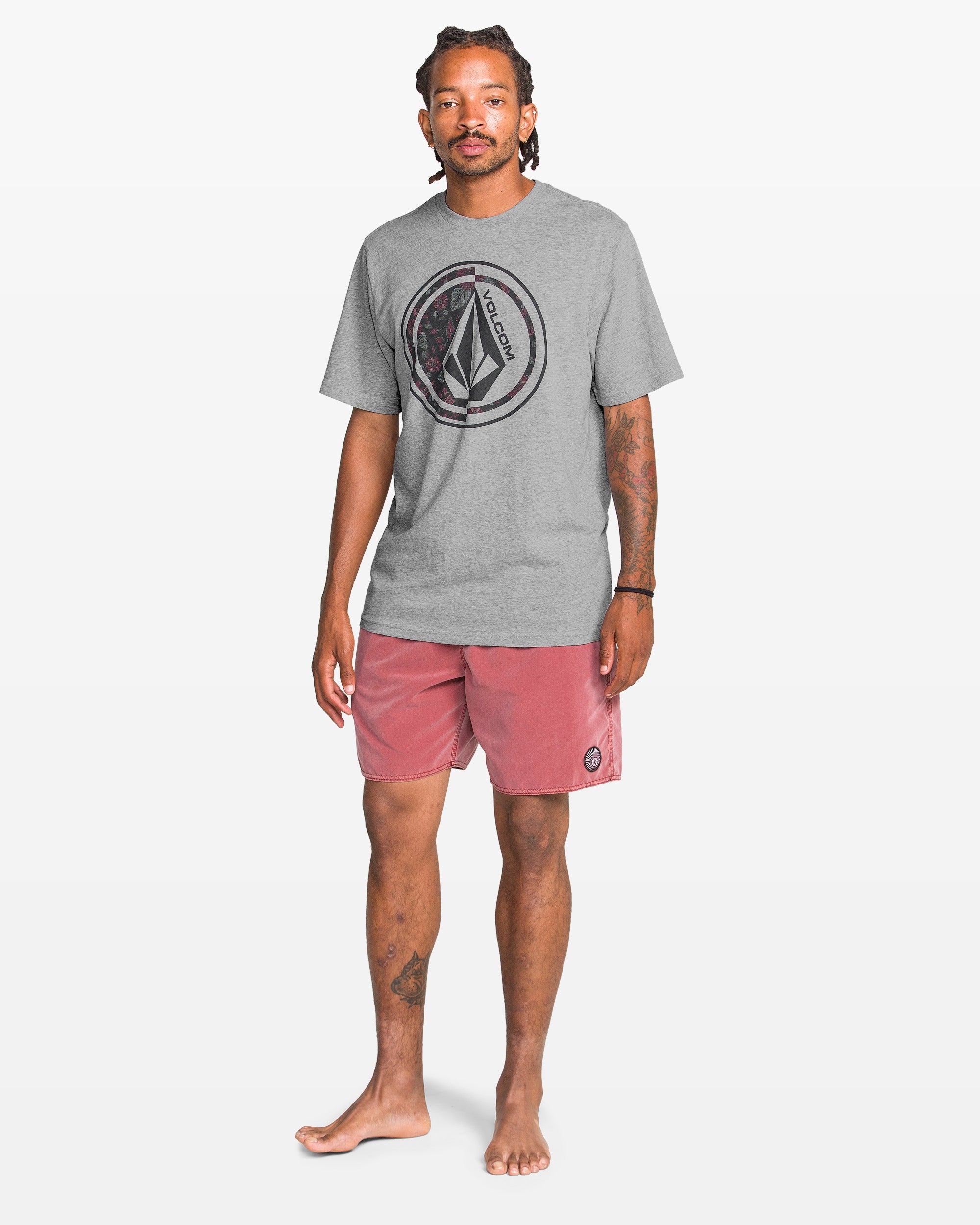 A man with braided hair and tattoos stands barefoot against a white background, wearing Volcom's Men's Coop Short Sleeve Tee in gray with a large logo, and dusty pink shorts featuring an emblem on the left leg.