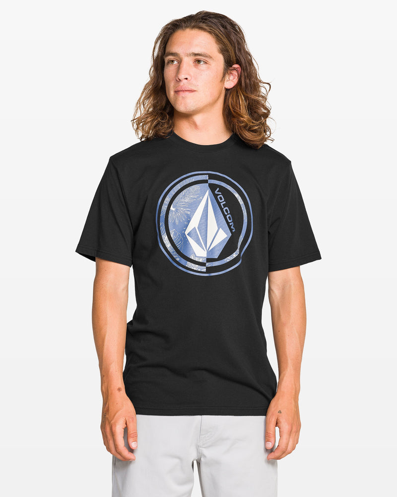 A person with long brown hair wears a black Volcom Men's Coop Short Sleeve Tee featuring a large circular graphic and light gray shorts, standing against a plain white background. The soft-hand screen print enhances the cotton-poly blend shirt’s comfort and style.