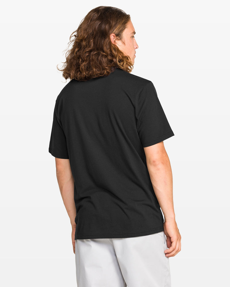 A person with long, wavy brown hair, seen from behind against a white background, is wearing a Volcom Men's Coop Short Sleeve Tee in plain black paired with light gray pants.