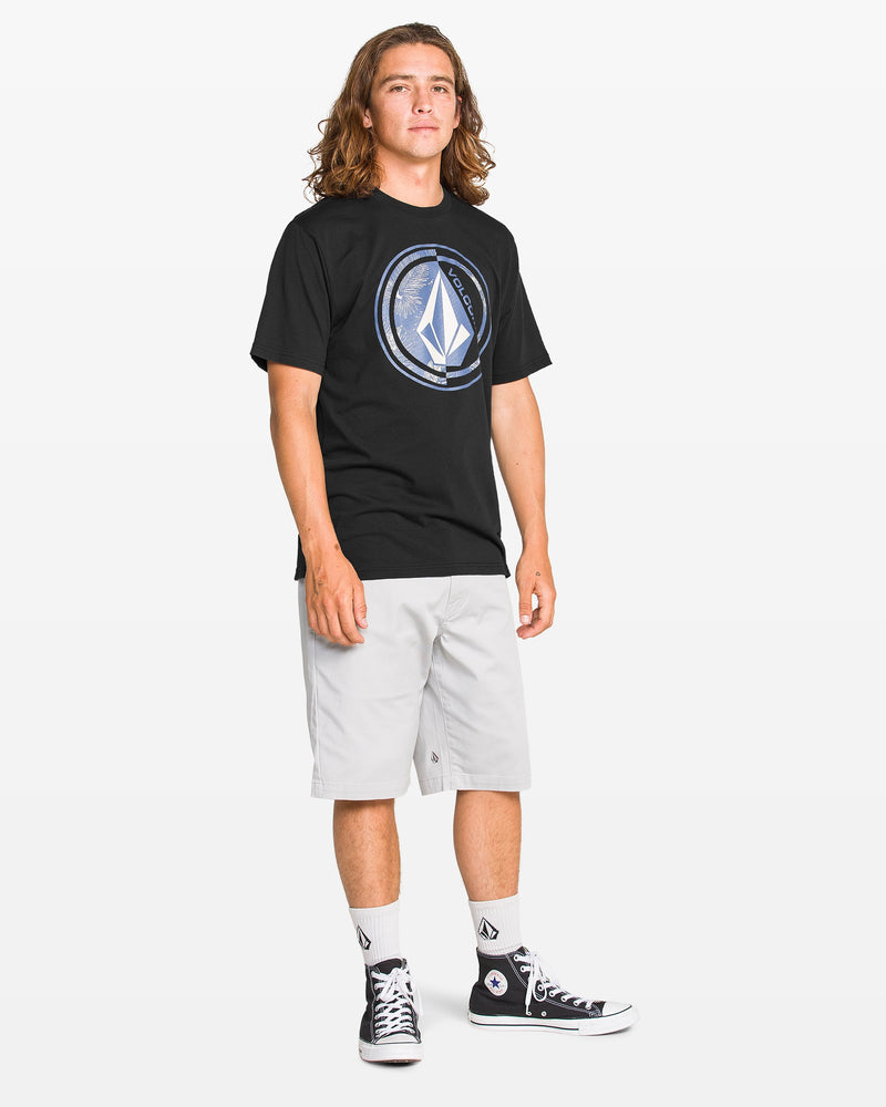 A young man with long hair wears the Volcom Men's Coop Short Sleeve Tee, light gray shorts, white socks, and black high-top sneakers, standing against a plain white background.