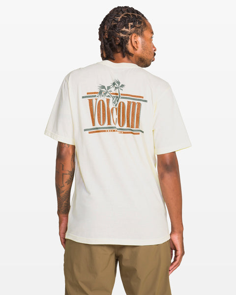 A man with short dreadlocks wears a cream Volcom Men's Breathe Deep Short Sleeve Tee featuring palm tree and logo graphics on the back, paired with light brown shorts, standing against a white background.