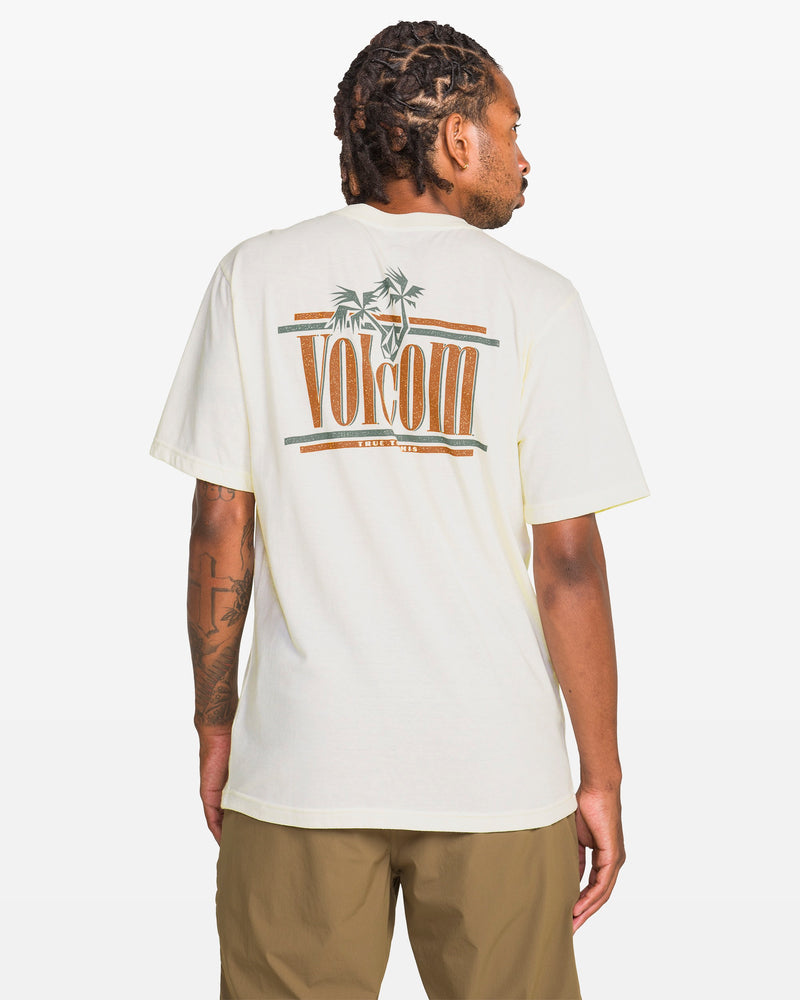 A man with short dreadlocks wears a cream Volcom Men's Breathe Deep Short Sleeve Tee featuring palm tree and logo graphics on the back, paired with light brown shorts, standing against a white background.