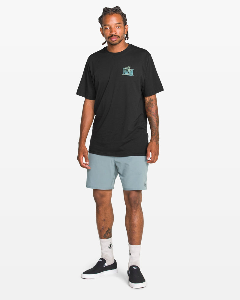 A man with braided hair stands against a white background wearing the Volcom Men's Breathe Deep Short Sleeve Tee in black with a small teal graphic, light blue shorts, white socks, and black slip-on sneakers.