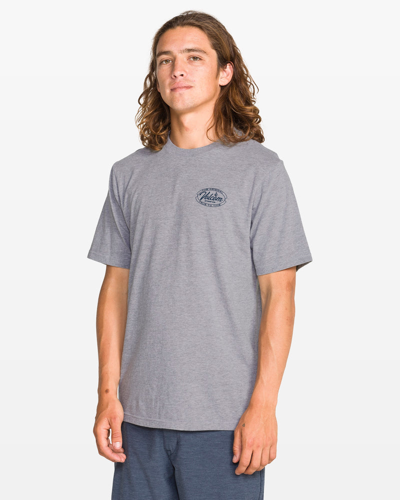 A young man with long brown hair stands confidently against a plain background, wearing the Volcom Men's Garaged Short Sleeve Tee in heather gray with a small black chest graphic and blue shorts, smiling slightly at the camera.