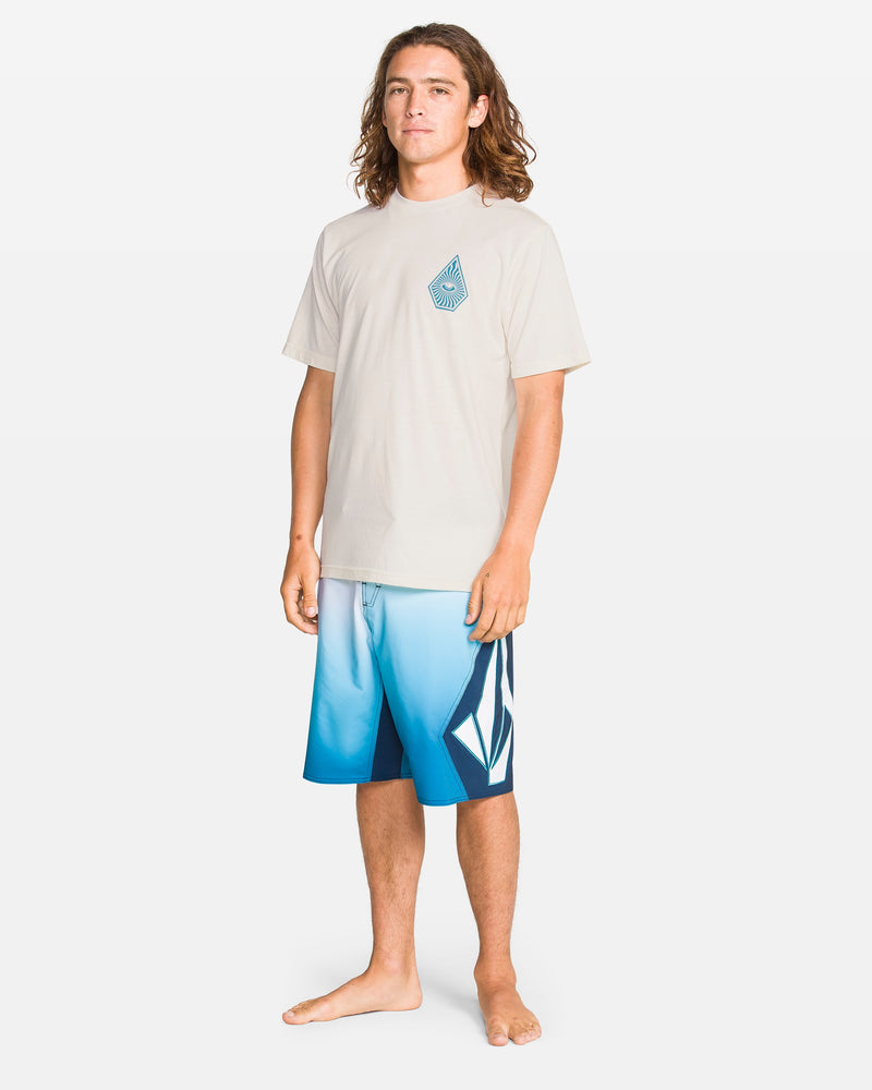 A young man with long brown hair stands barefoot, wearing the Volcom Men's All Seeing Short Sleeve Tee in Off White Heather, which has a small blue chest graphic, paired with modern-fit blue gradient board shorts featuring bold white and dark blue patterns.