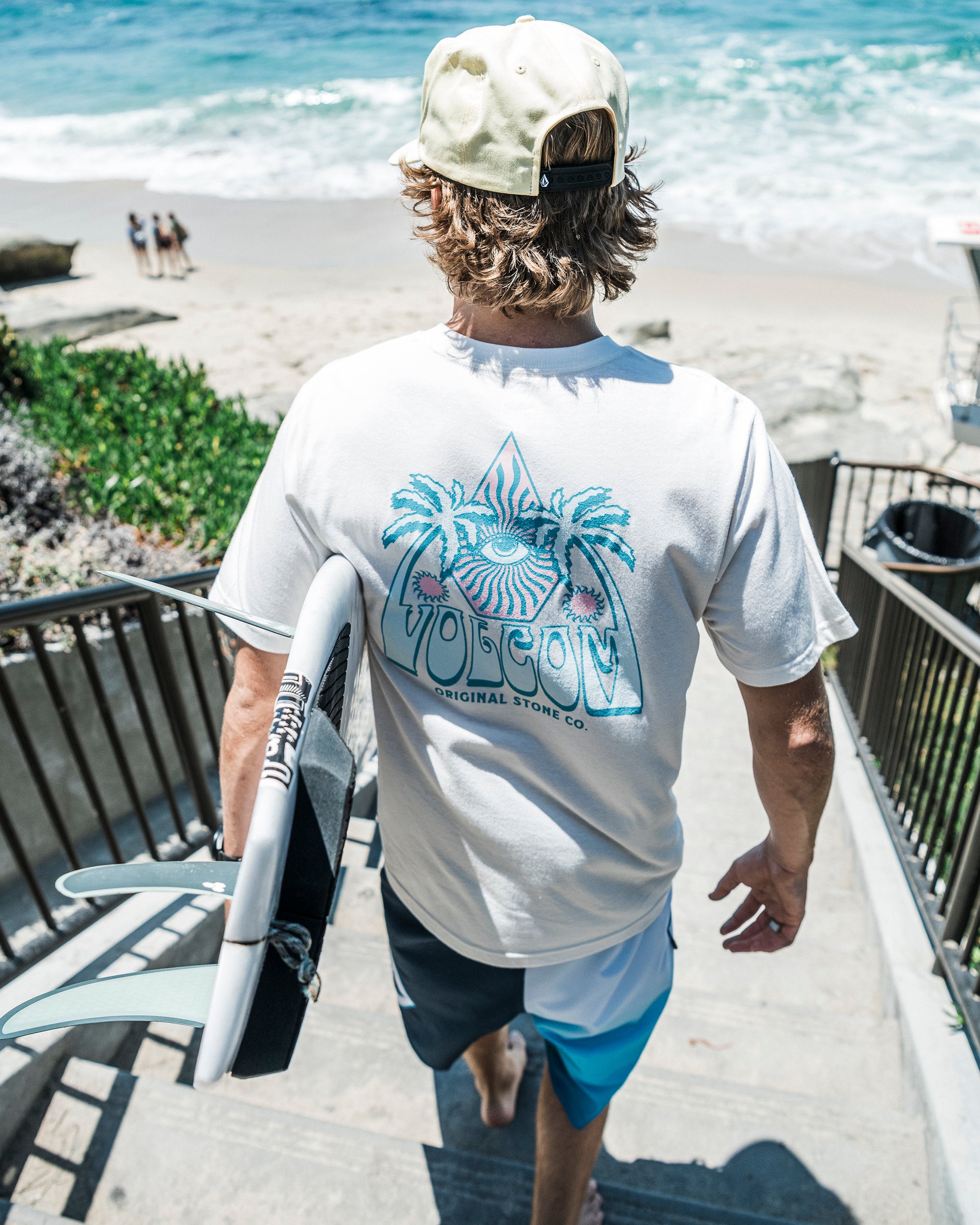 A man in a backwards cap and Volcom Men's All Seeing Short Sleeve Tee carries a surfboard down stairs toward a sandy beach and blue ocean on a sunny day.