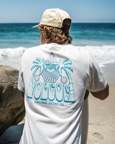 Wearing a Volcom Men's All Seeing Short Sleeve Tee in Off White Heather with a vibrant eye and palm tree graphic, plus a beige cap, someone stands on the sand facing the ocean. The tee is made of ring spun cotton and says "Volcom Original Stone Co.