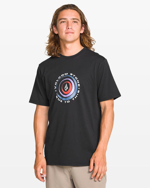A man with long brown hair wears a black Volcom Men's Enterprise Short Sleeve Tee in a modern fit, featuring a circular chest logo and text, paired with light pants, standing against a plain white background.