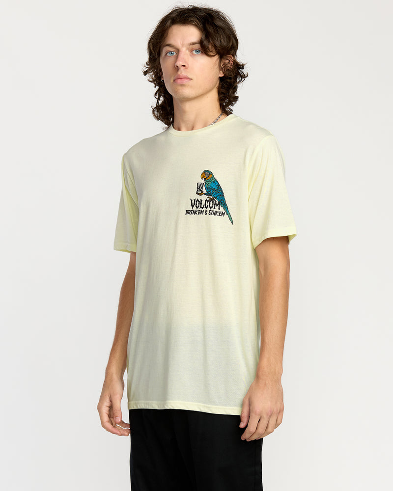 A young man with wavy brown hair wears the Volcom Men's Rambln Short Sleeve Tee, a relaxed fit light yellow cotton blend shirt featuring a colorful parrot graphic and chest text. He stands against a plain white background, looking slightly to the side.