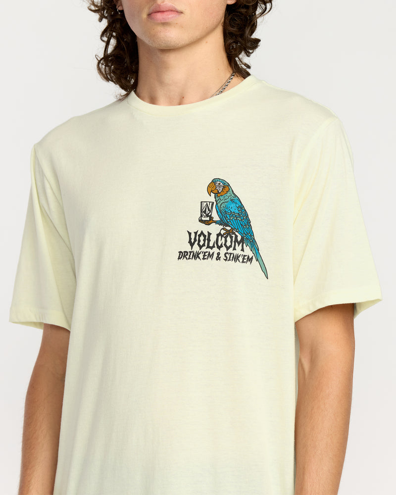 A person wears the Volcom Men's Rambln Short Sleeve Tee—a relaxed fit, light yellow cotton blend shirt featuring a colorful parrot holding a glass and the "Drink Em & Sink Em" graphic. The person's face is partially out of frame.