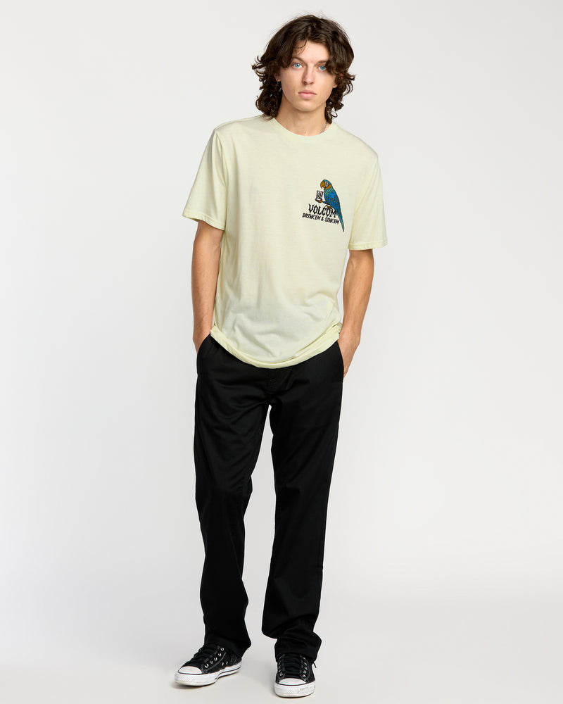A young man with wavy brown hair stands against a plain background, wearing the Volcom Men's Rambln Short Sleeve Tee. He pairs it with black pants and sneakers, hands in his pockets and a relaxed expression on his face.
