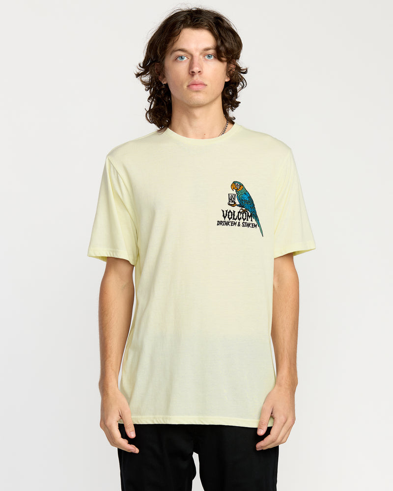 A young man with wavy brown hair stands forward, wearing the Volcom Men's Rambln Short Sleeve Tee—a relaxed fit, light yellow cotton blend shirt with a small parrot and text graphic on the left chest. He has a neutral expression and wears black pants.