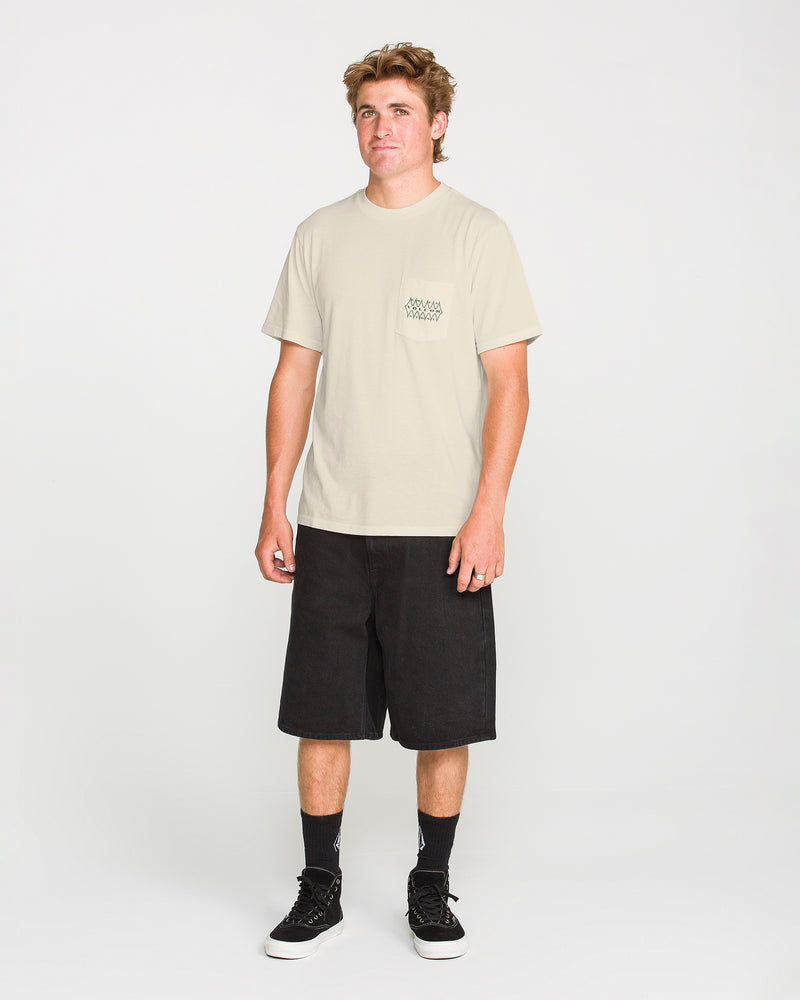 A person stands facing forward wearing the Volcom Men's Smokers Lounge Short Sleeve Tee in light beige, paired with black knee-length shorts, black crew socks, and black sneakers against a plain white background.