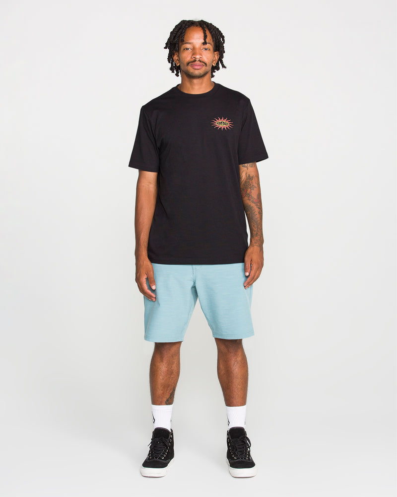 A man stands facing forward against a plain white background, wearing the Volcom Men's Spf91 Short Sleeve Tee—a medium-weight heather shirt with a small sun graphic—paired with light blue shorts, white socks, and black sneakers. The tee features a modern fit and is crafted from a soft ring spun cotton-poly blend for everyday comfort.