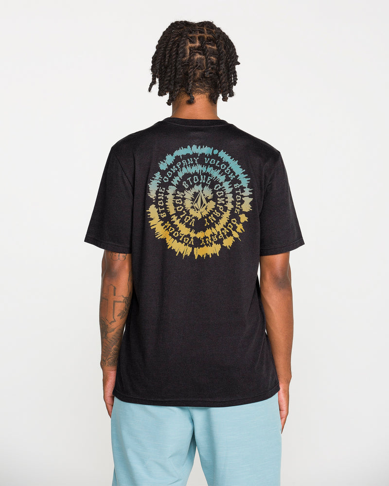A person with short dreadlocks wears the Volcom Men's Still Spiraling Short Sleeve Tee, a washed black t-shirt featuring a colorful circular graphic and wavy text created with a soft hand screen print on the back. They pair it with light blue shorts while standing against a plain white background.