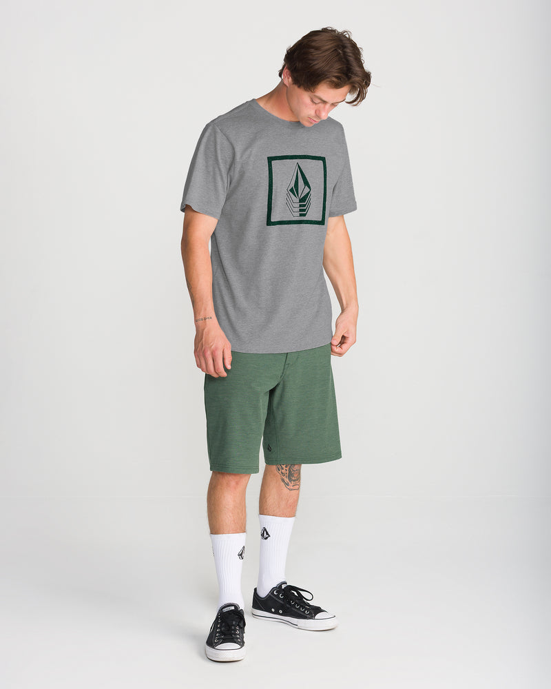 A person in a Volcom Men’s Quadratic Short Sleeve Tee (heather grey) with a geometric leaf logo, green shorts, white socks, and black sneakers stands against a plain white background, looking down.