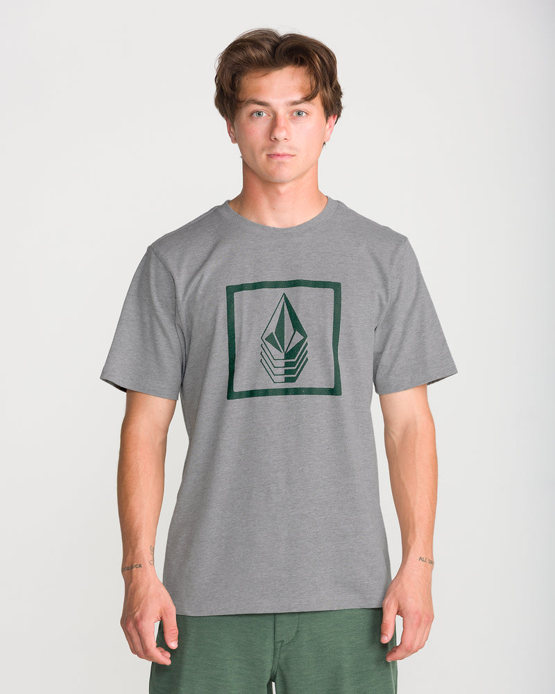 A young man wears the Volcom Men’s Quadratic Short Sleeve Tee in heather grey with a square geometric logo, modern fit, standing against a plain white background.