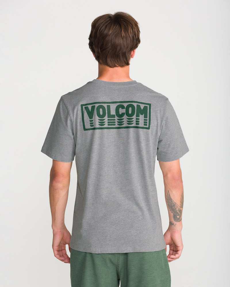 A person in a heather grey Volcom Men’s Quadratic Short Sleeve Tee with dark green back design faces away from the camera, paired with green shorts.