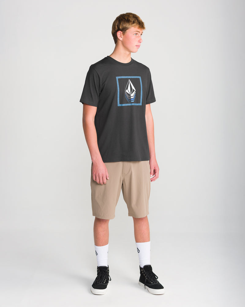 Teen boy wearing the Volcom Men’s Quadratic Short Sleeve Tee in washed black heather, beige knee-length shorts, white socks, and black sneakers stands in a studio with a plain light background, looking off to the side.