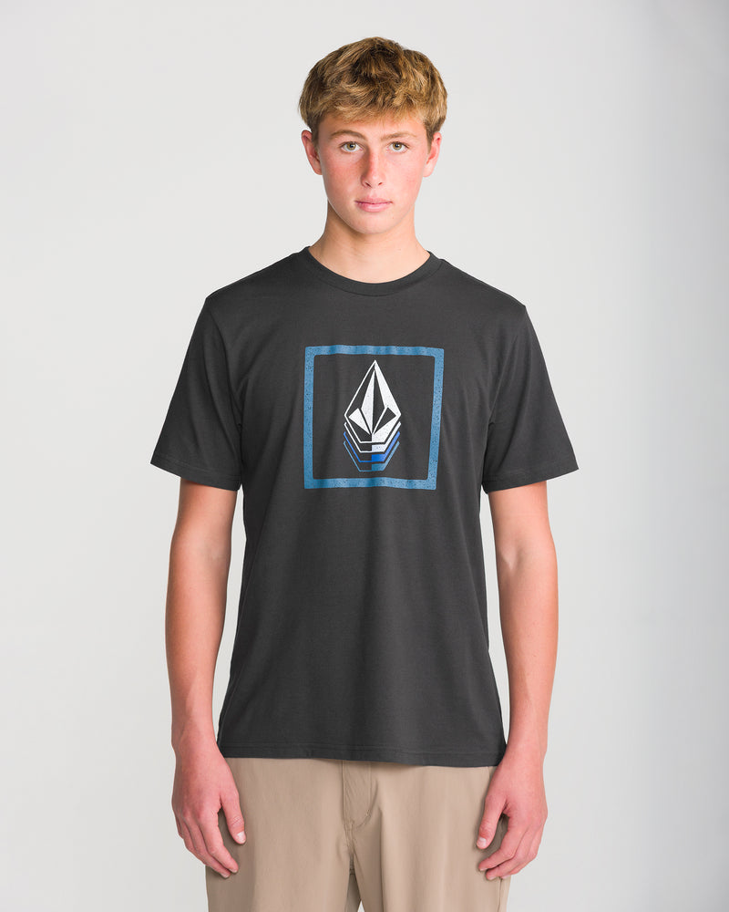 A teen boy with short blond hair wears the Volcom Men’s Quadratic Short Sleeve Tee in washed black heather with a geometric logo inside a blue square, paired with tan pants, against a plain light background.