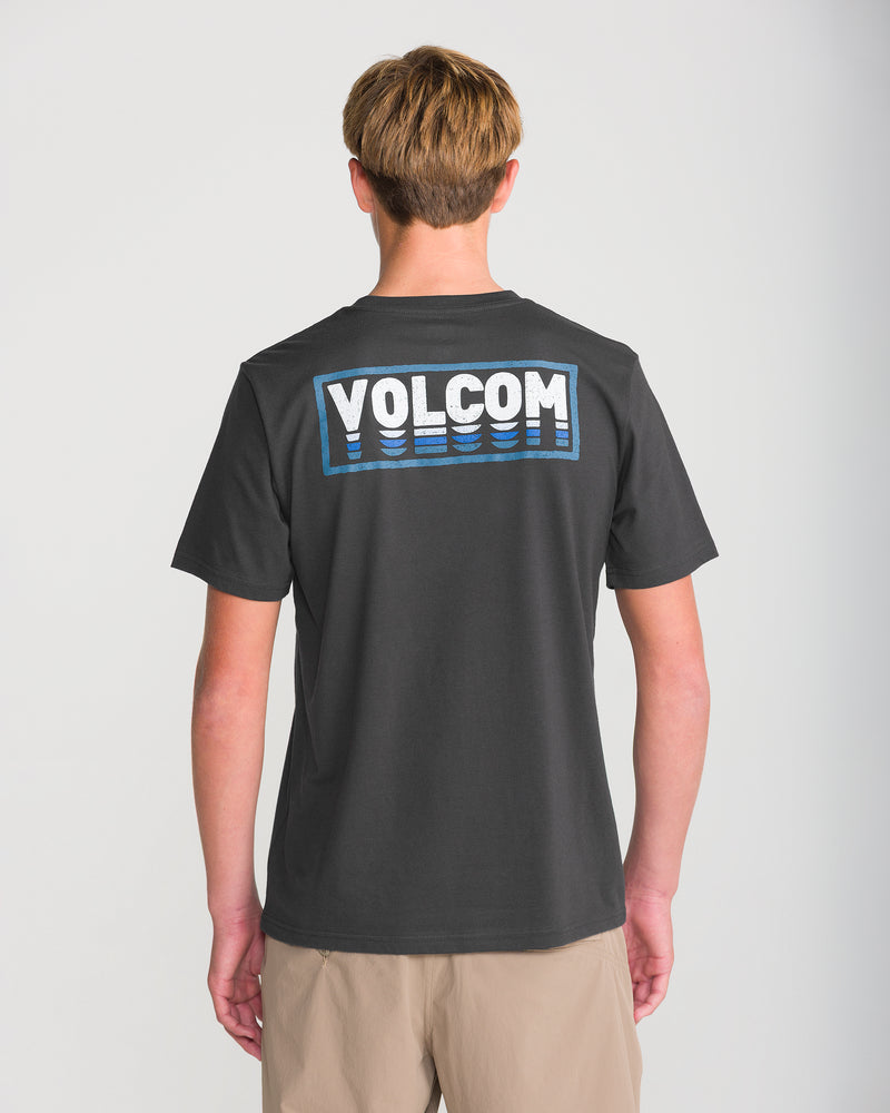 A person faces away, wearing the Men’s Quadratic Short Sleeve Tee by Volcom in washed black heather with white and blue VOLCOM print on the back, paired with light shorts against a plain, light background.